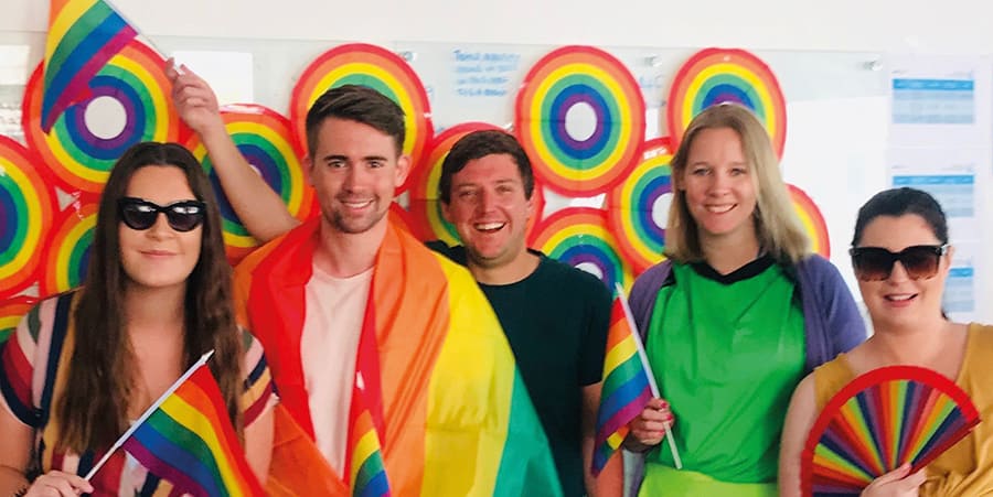 Five people smiling at the camera, waving rainbow coloured flags and folding fans