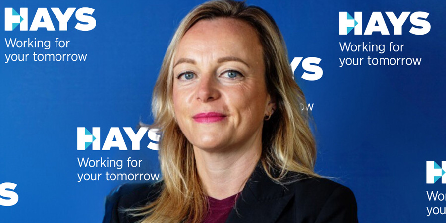 A woman looks into the camera, smiling softly. The background is blue, with the text Hays working for your tomorrow printed on the wall in white