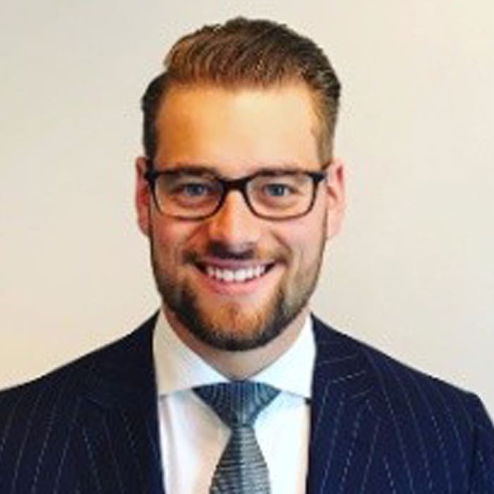 A young man wearing a suit and glasses smiles at the camera. The background is blank.